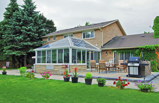 The exterior of a Georgian conservatory in the back of a home with an extended patio that has a table and grilling equipment.